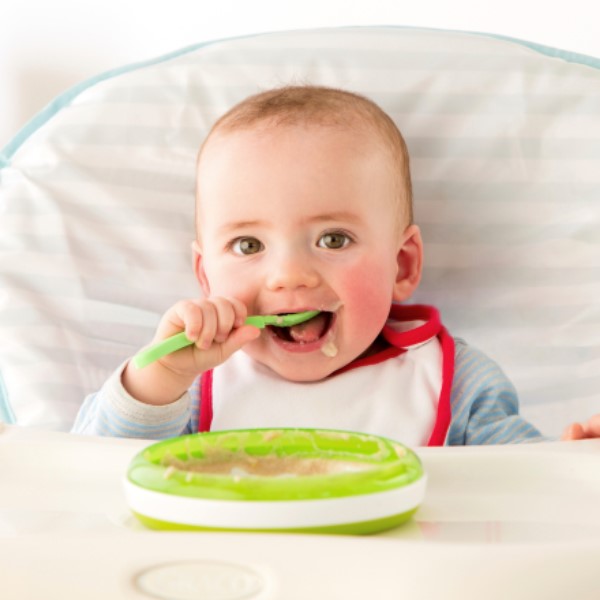 An infant in a high chair happliy feeding themself independently with a spoon