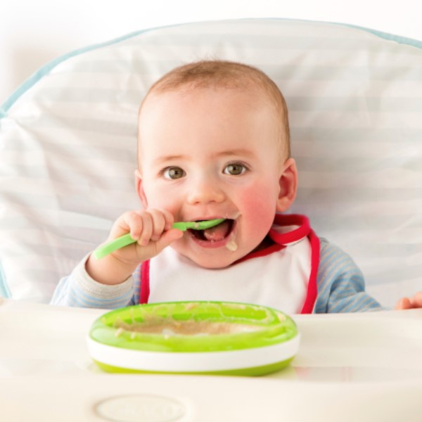 An infant sitting in a high chair happily feeding themself independently with a spoon